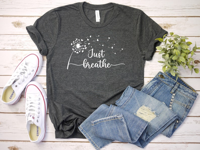 K&ouml;nnte beinhalten: Ein dunkelgraues T-Shirt mit einem wei&szlig;en L&ouml;wenzahnmuster und dem Text "Just breathe". Das Shirt liegt auf einer wei&szlig;en Holzoberfl&auml;che mit einem Paar wei&szlig;en Turnschuhen und einer blauen Jeans.