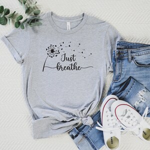K&ouml;nnte beinhalten: Ein graues T-Shirt mit einem schwarzen L&ouml;wenzahnmuster und dem Text "Just breathe".