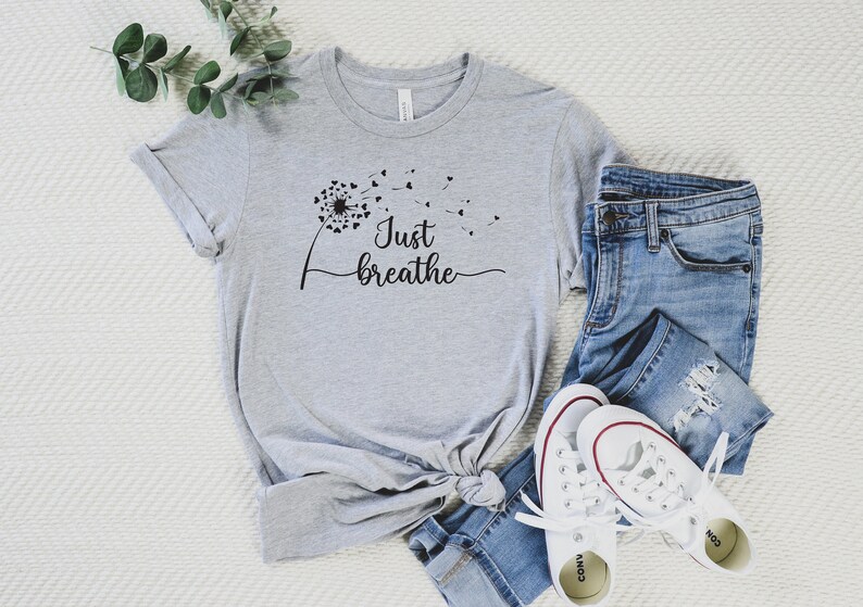 K&ouml;nnte beinhalten: Ein graues T-Shirt mit einem schwarzen L&ouml;wenzahnmuster und dem Text "Just breathe".