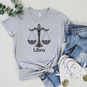 Op de afbeelding: Een grijs T-shirt met een zwart Weegschaal-sterrenbeeld en het woord "Weegschaal" erop gedrukt. Het shirt is gevouwen en ligt op een wit oppervlak met een blauwe spijkerbroek en witte sneakers.