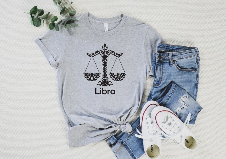 Op de afbeelding: Een grijs T-shirt met een zwart Weegschaal-sterrenbeeld en het woord "Weegschaal" erop gedrukt. Het shirt is gevouwen en ligt op een wit oppervlak met een blauwe spijkerbroek en witte sneakers.
