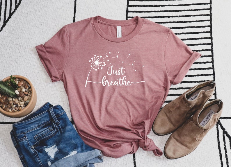 K&ouml;nnte beinhalten: Ein mauvefarbenes T-Shirt mit einem wei&szlig;en L&ouml;wenzahnmuster und dem Text "Just Breathe" in einer kursivschrift.