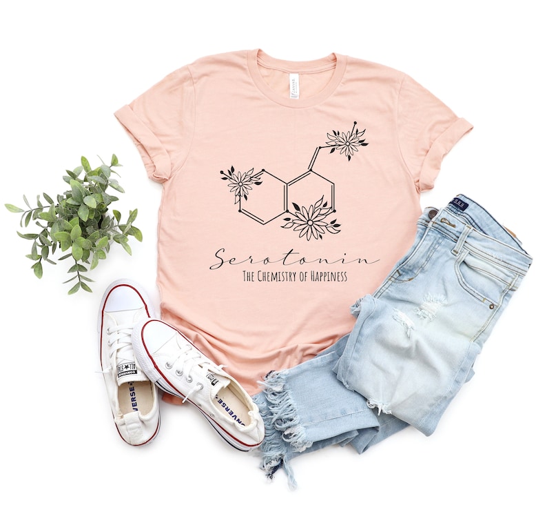 Pu&ograve; includere: Una maglietta rosa con una grafica nera di una molecola di serotonina con fiori. Il testo "Serotonina La chimica della felicit&agrave;" &egrave; stampato sotto la grafica.