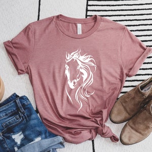 May include: A mauve t-shirt with a white silhouette of a horse's head on the front. The shirt is laid flat on a white and black striped rug with a pair of brown suede ankle boots and a pair of blue denim shorts.