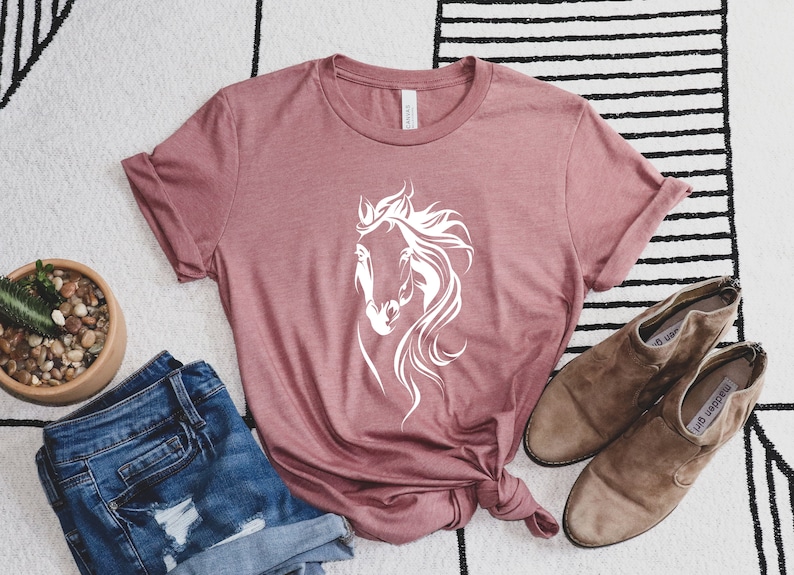 May include: A mauve t-shirt with a white silhouette of a horse's head on the front. The shirt is laid flat on a white and black striped rug with a pair of brown suede ankle boots and a pair of blue denim shorts.