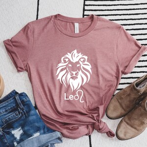 Może przedstawiać: R&oacute;żowy t-shirt z nadrukiem białego lwa i napisem "Leo" w kolorze białym.