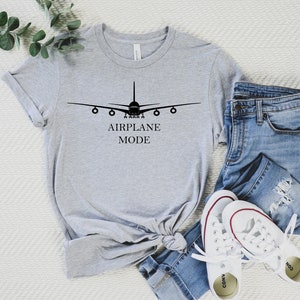 May include: A gray t-shirt with a black airplane graphic and the text "AIRPLANE MODE". The shirt is laid out on a white surface with a pair of blue jeans and white sneakers.