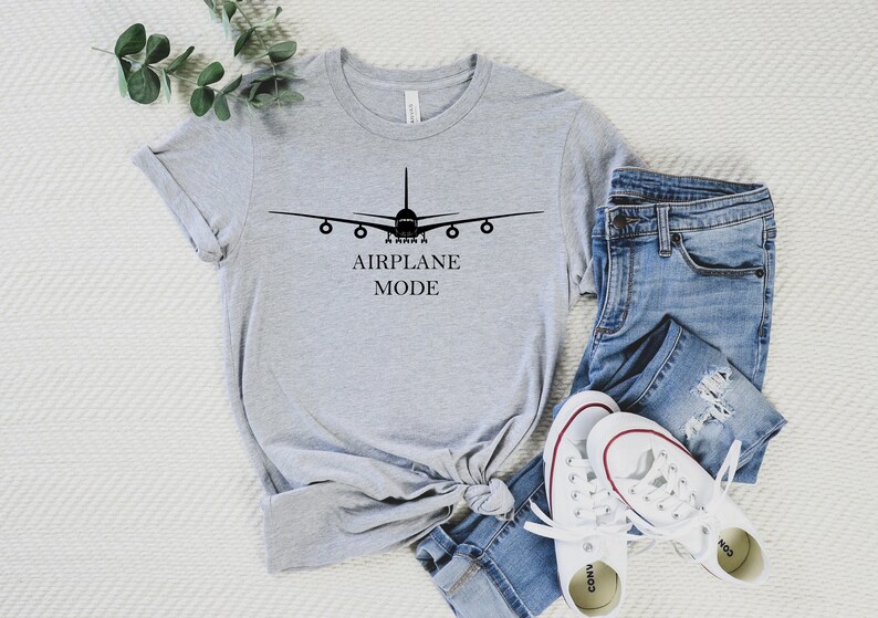 May include: A gray t-shirt with a black airplane graphic and the text "AIRPLANE MODE". The shirt is laid out on a white surface with a pair of blue jeans and white sneakers.