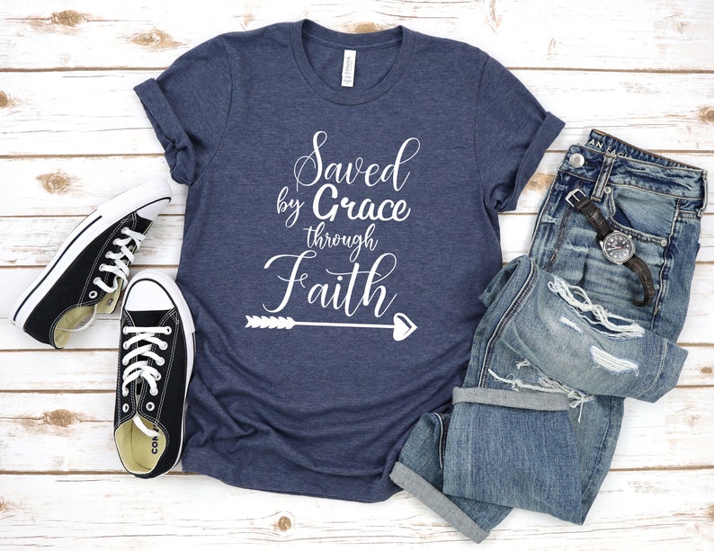 K&ouml;nnte beinhalten: Ein marineblaues T-Shirt mit dem Text "Saved by Grace through Faith" in wei&szlig;en Buchstaben. Das Shirt hat au&szlig;erdem einen wei&szlig;en Pfeil mit einem Herz am Ende. Das Shirt liegt auf einer wei&szlig;en Holzoberfl&auml;che mit einem Paar schwarzen Canvas-Schuhen und einer blauen Jeans.