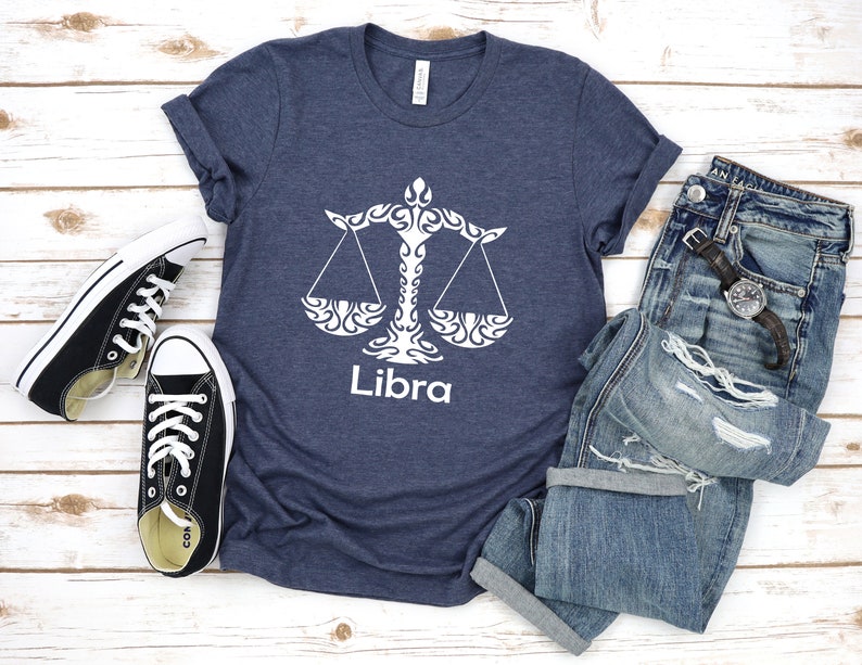 Op de afbeelding: Een marineblauw t-shirt met een wit Libra-sterrenbeeld en het woord "Libra" erop gedrukt. Het shirt ligt plat op een wit houten oppervlak met een paar zwarte Converse sneakers en een blauwe jeans.