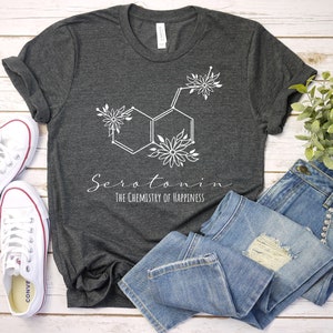 Pu&ograve; includere: Una maglietta grigio scuro con un design floreale bianco della struttura chimica della serotonina. Il testo "Serotonina La chimica della felicit&agrave;" &egrave; stampato sotto il design.