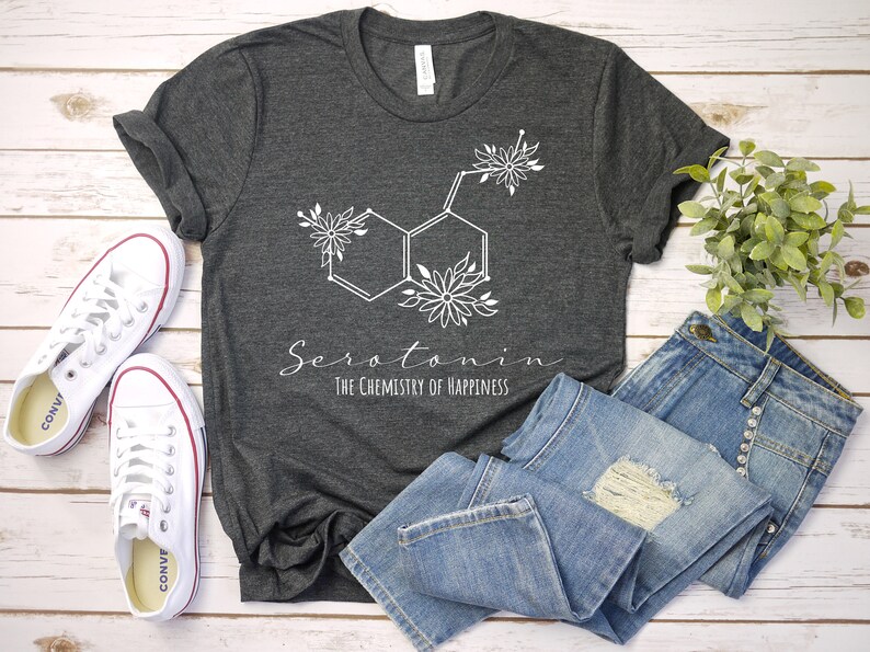 Pu&ograve; includere: Una maglietta grigio scuro con un design floreale bianco della struttura chimica della serotonina. Il testo "Serotonina La chimica della felicit&agrave;" &egrave; stampato sotto il design.