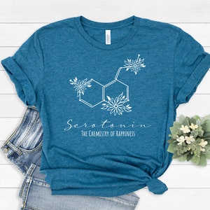 Pu&ograve; includere: Maglietta blu turchese con una grafica di molecola di serotonina floreale bianca e il testo "Serotonina La chimica della felicit&agrave;".