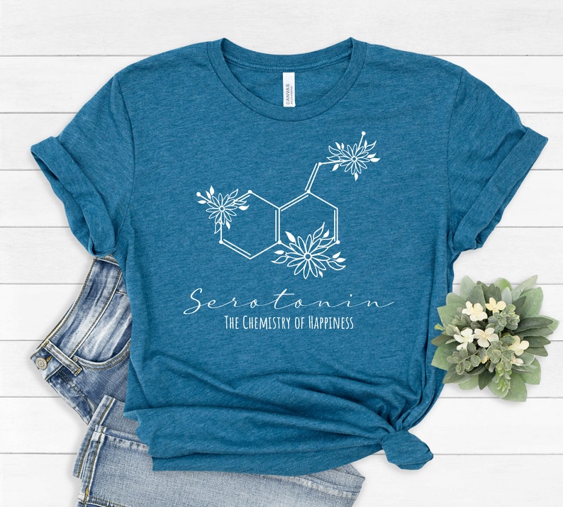 Pu&ograve; includere: Maglietta blu turchese con una grafica di molecola di serotonina floreale bianca e il testo "Serotonina La chimica della felicit&agrave;".