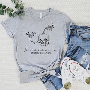 Pu&ograve; includere: Una maglietta grigia con una grafica nera di una molecola di serotonina con accenti floreali. Il testo "Serotonina La chimica della felicit&agrave;" &egrave; stampato sotto la molecola.