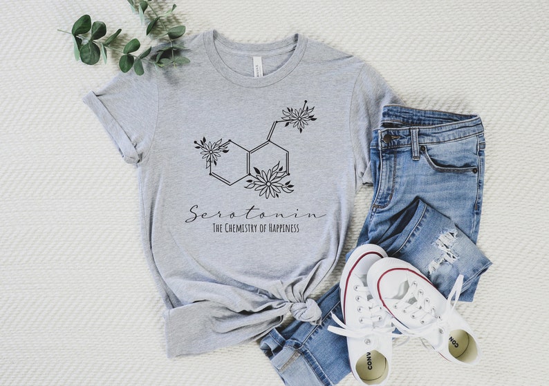 Pu&ograve; includere: Una maglietta grigia con una grafica nera di una molecola di serotonina con accenti floreali. Il testo "Serotonina La chimica della felicit&agrave;" &egrave; stampato sotto la molecola.
