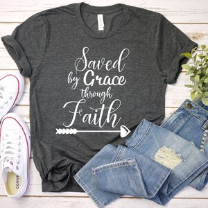 K&ouml;nnte beinhalten: Ein dunkelgraues T-Shirt mit dem Text "Saved by Grace through Faith" in wei&szlig;en Buchstaben mit einem Pfeil- und Herzdesign. Das Shirt liegt flach auf einer wei&szlig;en Holzoberfl&auml;che mit einem Paar wei&szlig;en Turnschuhen und einer blauen Jeans.