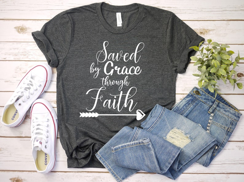K&ouml;nnte beinhalten: Ein dunkelgraues T-Shirt mit dem Text "Saved by Grace through Faith" in wei&szlig;en Buchstaben mit einem Pfeil- und Herzdesign. Das Shirt liegt flach auf einer wei&szlig;en Holzoberfl&auml;che mit einem Paar wei&szlig;en Turnschuhen und einer blauen Jeans.
