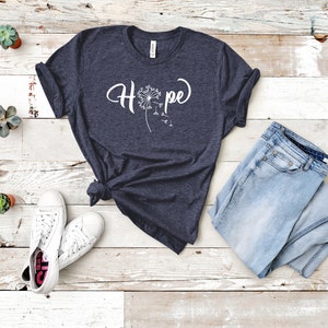 May include: A dark blue t-shirt with a white dandelion graphic and the word "Hope" printed on the front. The shirt is laid flat on a white wooden surface with a pair of white sneakers and blue jeans.