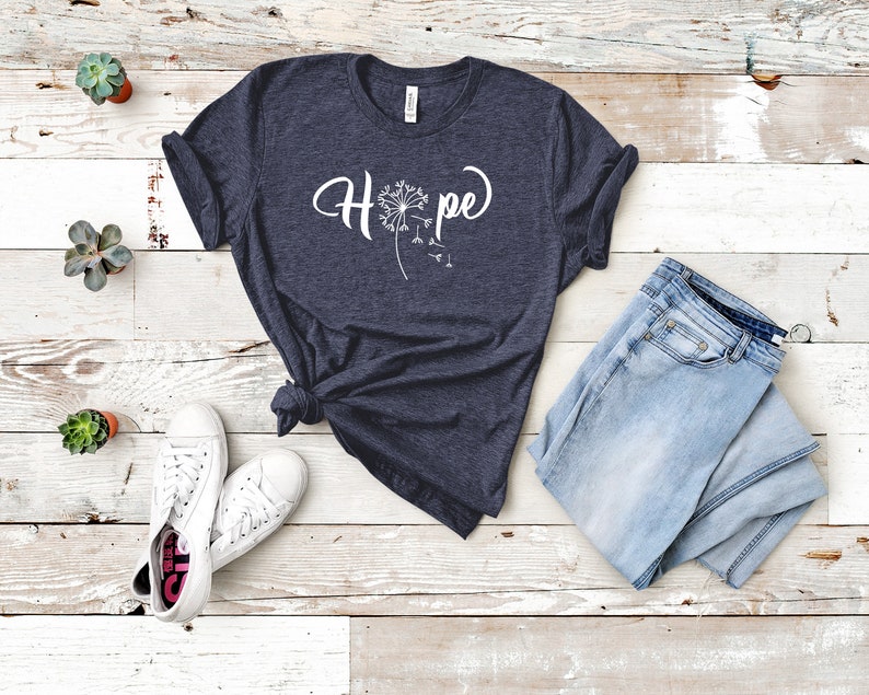 May include: A dark blue t-shirt with a white dandelion graphic and the word "Hope" printed on the front. The shirt is laid flat on a white wooden surface with a pair of white sneakers and blue jeans.