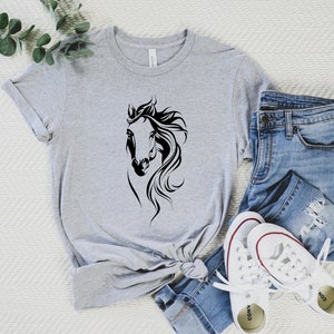 May include: A gray t-shirt with a black silhouette of a horse's head. The shirt is styled with blue jeans and white sneakers.