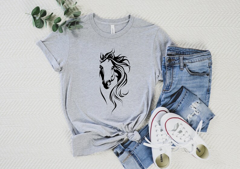 May include: A gray t-shirt with a black silhouette of a horse's head. The shirt is styled with blue jeans and white sneakers.