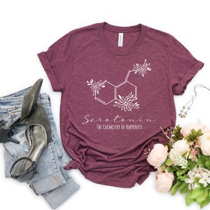Pu&ograve; includere: Una maglietta color granata con una grafica bianca della struttura chimica della serotonina con accenti floreali. Il testo "Serotonina La chimica della felicit&agrave;" &egrave; stampato sotto la grafica.