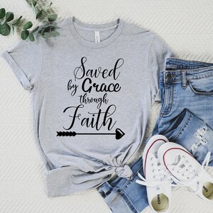 K&ouml;nnte beinhalten: Ein graues T-Shirt mit dem Text "Saved by Grace through Faith" in schwarzer Kursivschrift mit einem Pfeil- und Herzdesign.