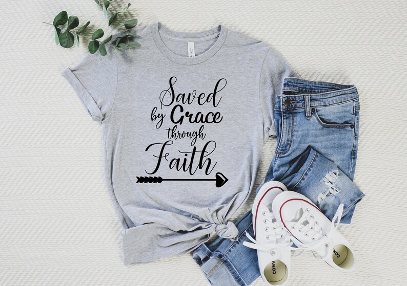 K&ouml;nnte beinhalten: Ein graues T-Shirt mit dem Text "Saved by Grace through Faith" in schwarzer Kursivschrift mit einem Pfeil- und Herzdesign.