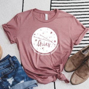 Pu&ograve; includere: Una maglietta rosa con un design circolare bianco che presenta il segno zodiacale Ariete e le parole "coraggioso generoso passionale".