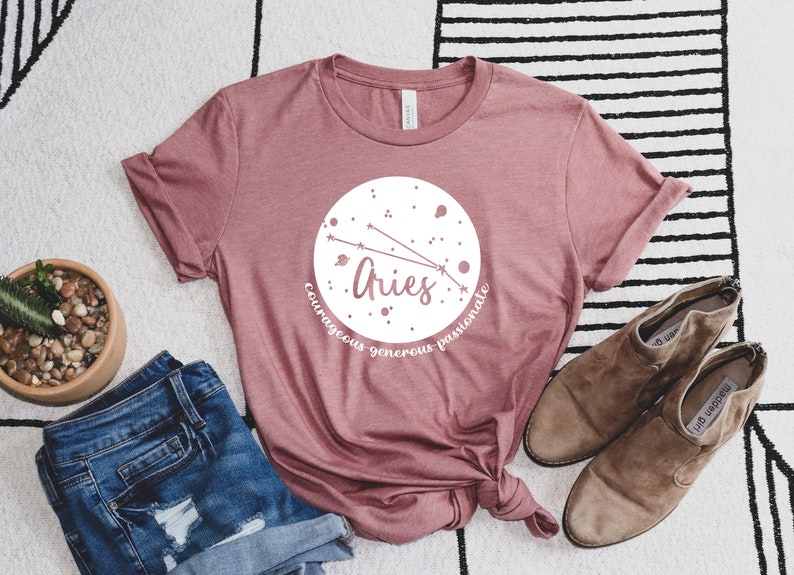 Pu&ograve; includere: Una maglietta rosa con un design circolare bianco che presenta il segno zodiacale Ariete e le parole "coraggioso generoso passionale".
