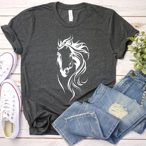 May include: A dark gray t-shirt with a white silhouette of a horse's head. The shirt is laid flat on a white wooden surface with a pair of white sneakers and a pair of blue jeans.