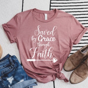 K&ouml;nnte beinhalten: Ein mauvefarbenes T-Shirt mit dem Text "Saved by Grace through Faith" in wei&szlig;en Buchstaben mit einem Pfeil- und Herzdesign.