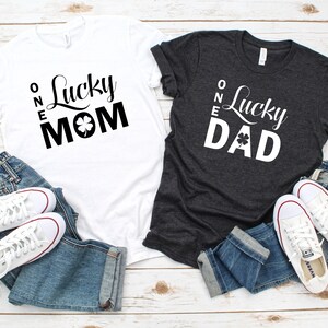May include: Two t-shirts with the text "One Lucky Mom" and "One Lucky Dad" printed on them. The shirts are white and dark grey, and each has a shamrock graphic.