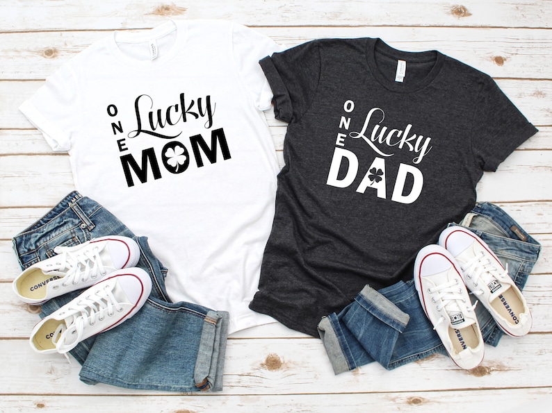 May include: Two t-shirts with the text "One Lucky Mom" and "One Lucky Dad" printed on them. The shirts are white and dark grey, and each has a shamrock graphic.