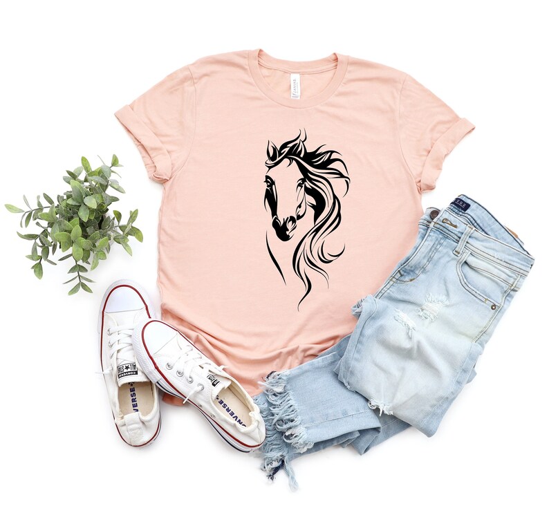 May include: A pink t-shirt with a black silhouette of a horse's head. The shirt is laying on a white surface with a pair of white sneakers and blue jeans.