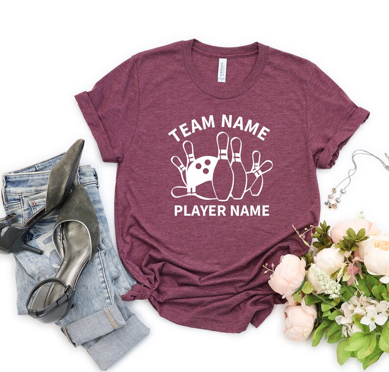 May include: Burgundy t-shirt with white text that reads "TEAM NAME" and "PLAYER NAME" with a graphic of bowling pins and a bowling ball.