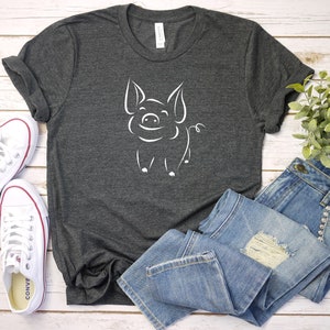 May include: A dark gray t-shirt with a white outline of a smiling pig on the front. The shirt is laid out on a white wooden surface with a pair of white sneakers, blue jeans, and a small potted plant.