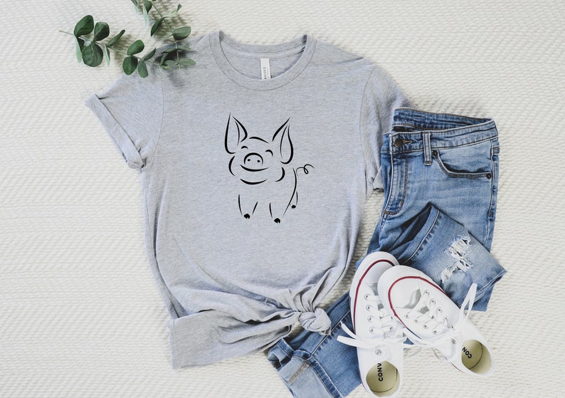 May include: A gray t-shirt with a black line drawing of a smiling pig on the front. The shirt is paired with blue jeans and white sneakers.