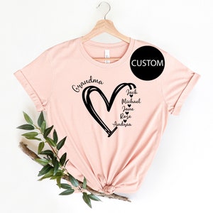 K&ouml;nnte beinhalten: Ein pinkfarbenes T-Shirt mit einem schwarzen Herzdesign und dem Wort "Grandma" dar&uuml;ber. Im Herzen stehen die Namen Jack, Michael, Jane, Rose und Andrea.