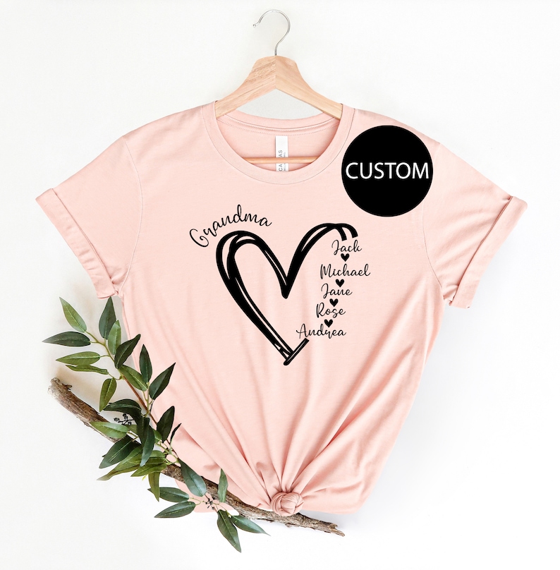K&ouml;nnte beinhalten: Ein pinkfarbenes T-Shirt mit einem schwarzen Herzdesign und dem Wort "Grandma" dar&uuml;ber. Im Herzen stehen die Namen Jack, Michael, Jane, Rose und Andrea.