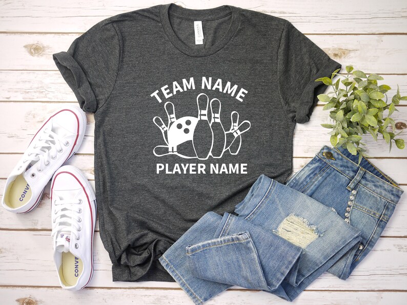 May include: Gray t-shirt with a white graphic of bowling pins and a bowling ball. The text "TEAM NAME" is above the graphic and "PLAYER NAME" is below the graphic.