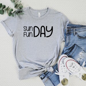 K&ouml;nnte beinhalten: Ein graues T-Shirt mit dem Text "Sun Fun Day" in schwarzen Buchstaben. Das Shirt ist gefaltet und wird mit einer blauen Jeans und wei&szlig;en Turnschuhen pr&auml;sentiert.