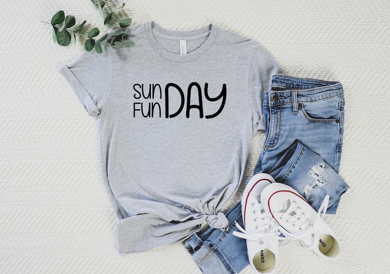 K&ouml;nnte beinhalten: Ein graues T-Shirt mit dem Text "Sun Fun Day" in schwarzen Buchstaben. Das Shirt ist gefaltet und wird mit einer blauen Jeans und wei&szlig;en Turnschuhen pr&auml;sentiert.