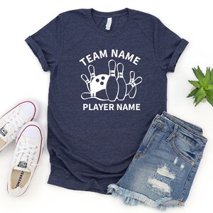 May include: A navy blue t-shirt with white text that reads "Team Name" and "Player Name". A white bowling pin graphic is centered on the shirt.