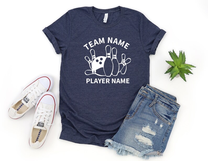 May include: A navy blue t-shirt with white text that reads "Team Name" and "Player Name". A white bowling pin graphic is centered on the shirt.