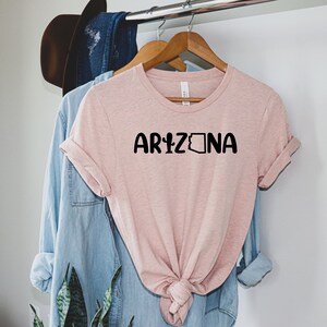 May include: A pink t-shirt with the text "ARIZONA" in black lettering. The text includes a cactus and the outline of the state of Arizona.