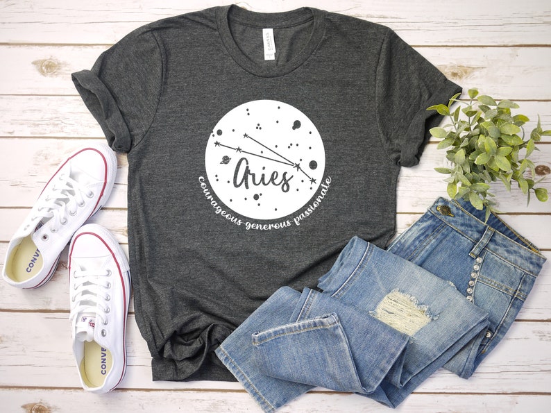 Pu&ograve; includere: Una maglietta grigio scuro con un design circolare bianco che presenta la costellazione zodiacale dell'Ariete. Il cerchio include anche la parola "Ariete" e le parole "coraggioso, generoso, passionale".