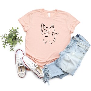 May include: A light pink t-shirt with a black line drawing of a pig on the front. The pig is smiling and has a curly tail. The shirt is paired with blue jeans and white sneakers.