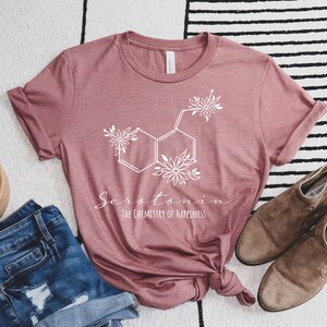 Pu&ograve; includere: Una maglietta color malva con una grafica bianca di una molecola di serotonina con accenti floreali. Il testo "Serotonina La chimica della felicit&agrave;" &egrave; stampato sotto la molecola.